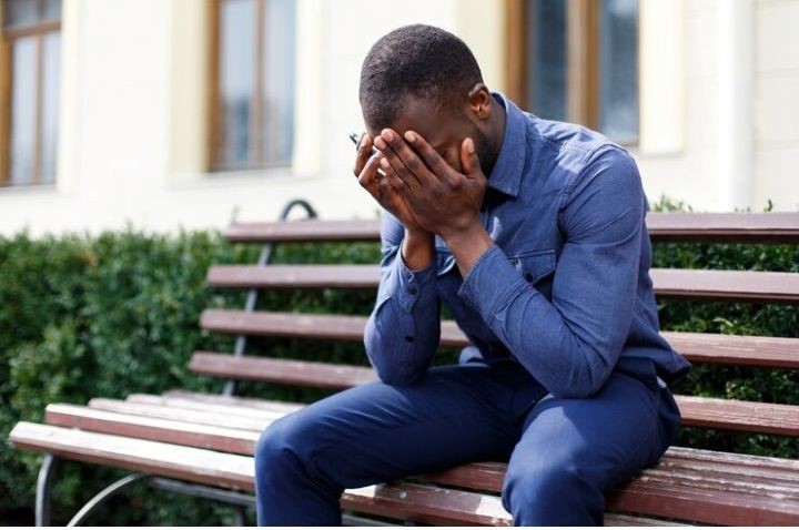 African man sitting outdoors on a wooden bench feeling sad due to his infertility