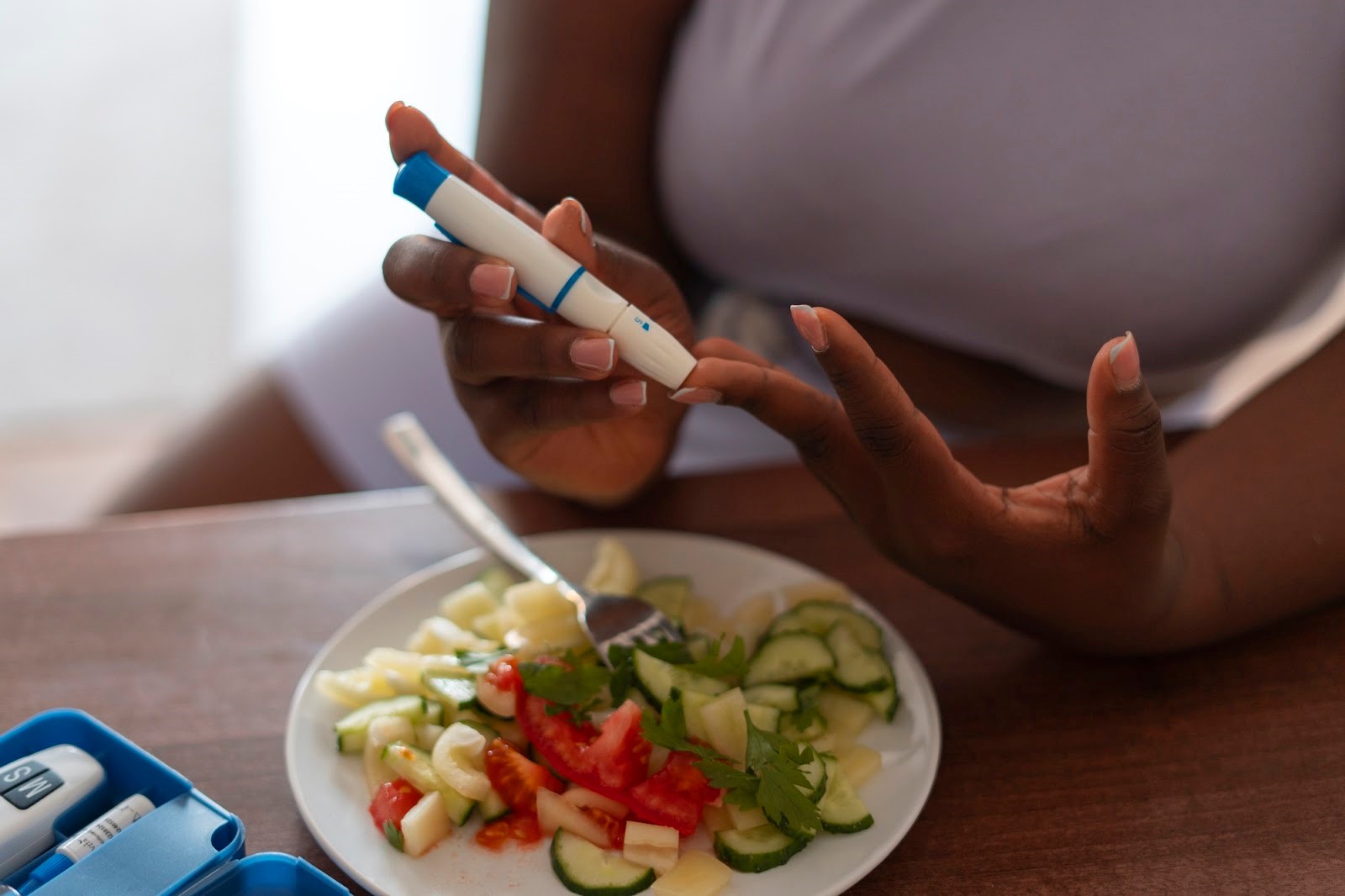 A black woman seated at a table with a plate of vegetable salad and glucose test kit, doing a finger prick to check her blood glucose level.