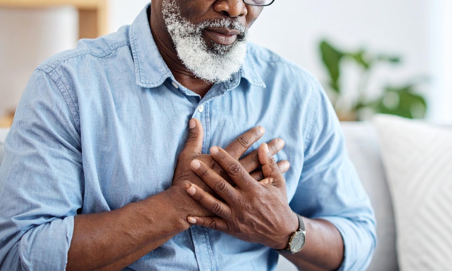 A professional looking African male wearing a casual blue shirt holding his left chest with both hands in apparent distress from sudden pain.