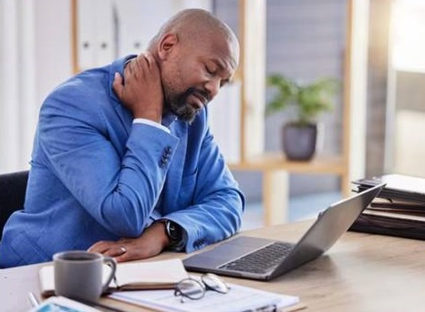 Exhausted and fatigued black man seated at a table before a laptop device holding his neck in pain in sign of burnout