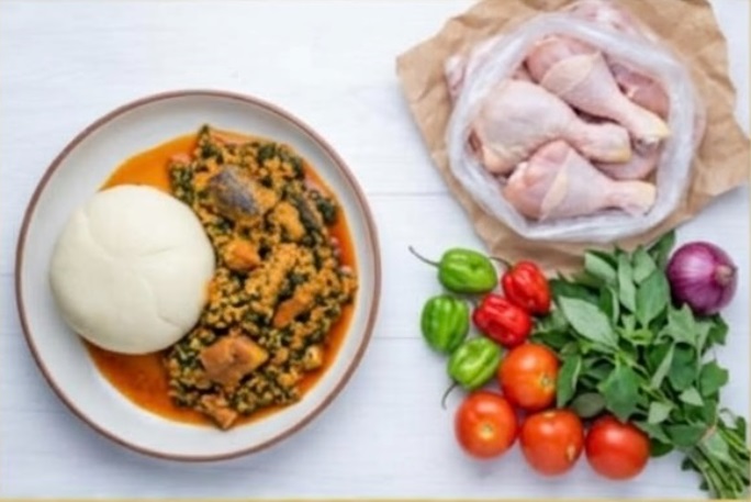 Photo of a top view of fresh vegetables such as pepper, carrot, onions, and chicken on a light desk kept separately from a plate of swalloq and vegetable stew.