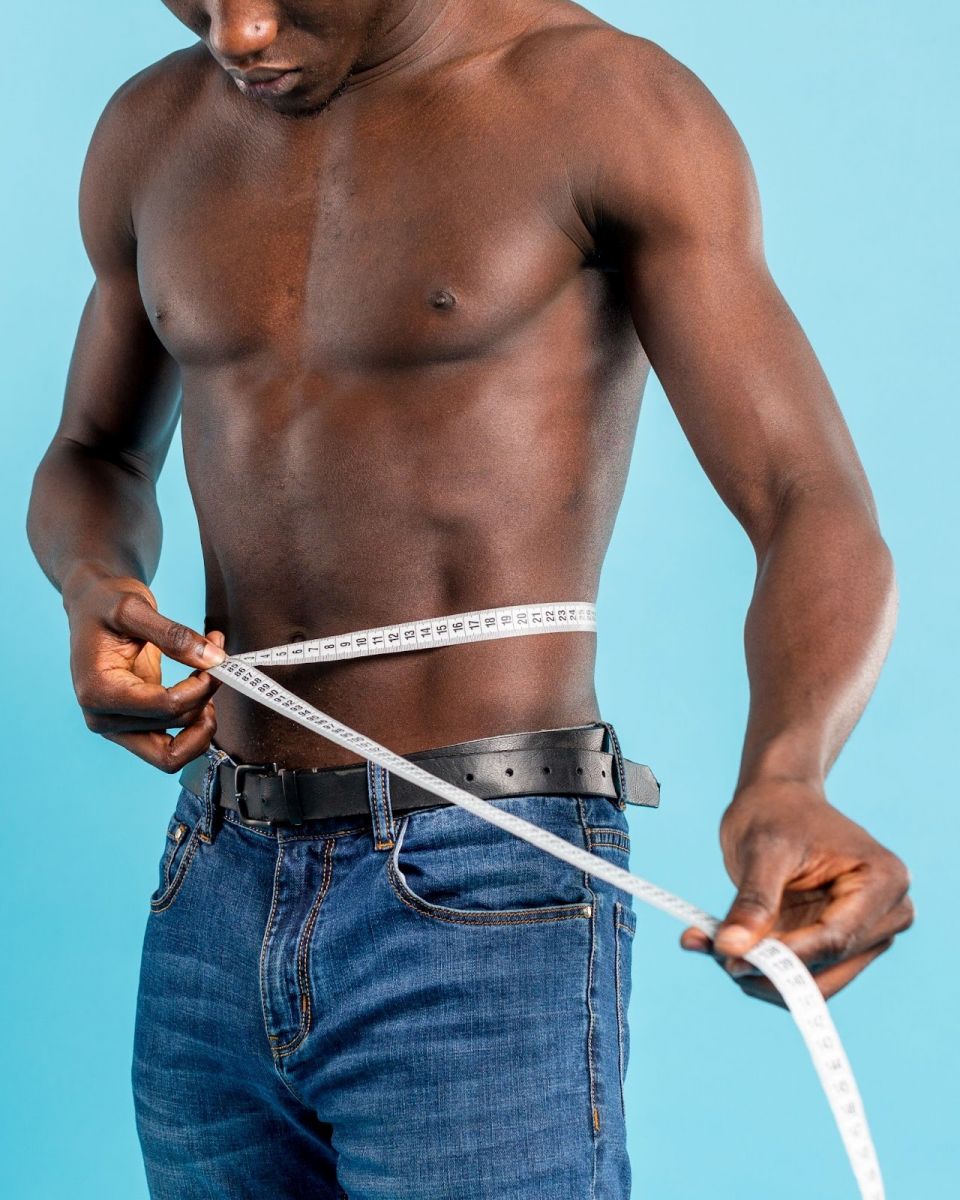 Black man measuring his waist with a tape measure against a blue background.