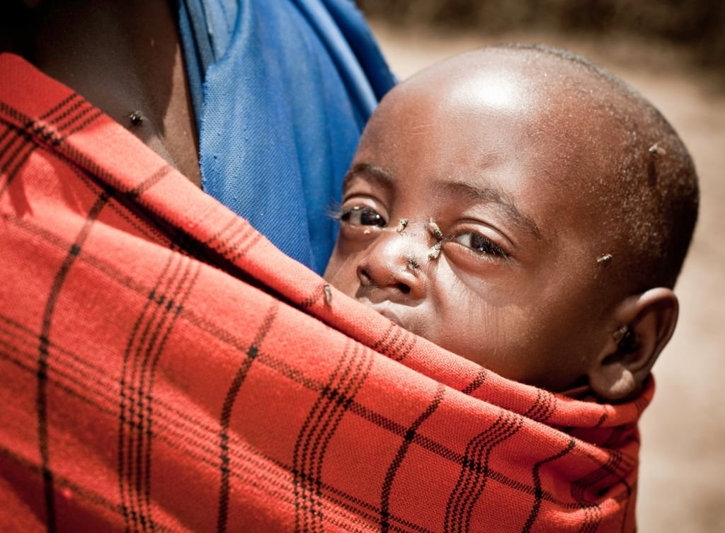 African mother backing a toddler with flies perching on his eyes and face