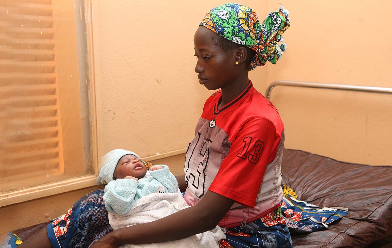 An adolescent African female sitting on a hospital bed and holding her newborn baby