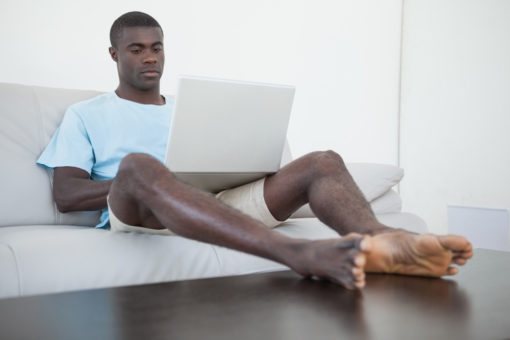 A young black man seated on a sofa with a laptop computer on the lap and with his feet up on a centre table across him.