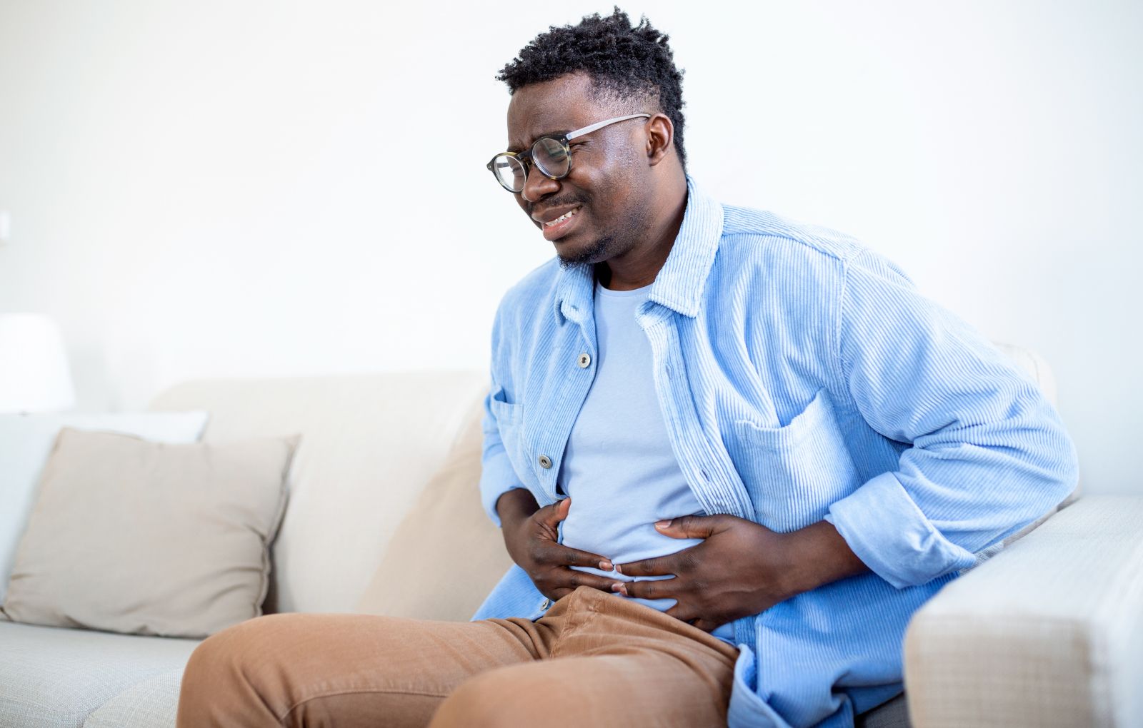 Young african sitting on a sofa and holding his belly in pain and obvious distress