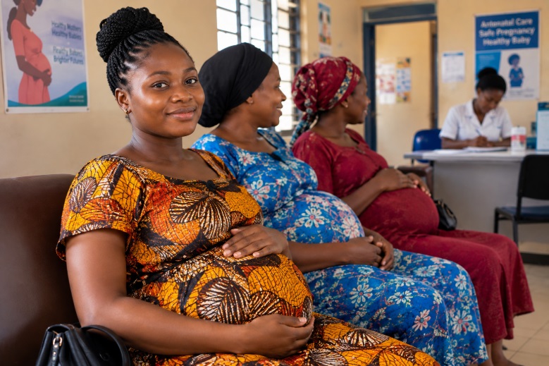 Image of three pregnant women sitting and awaiting care in an antenatal clinic with a nurse seated at a table in the far background. 