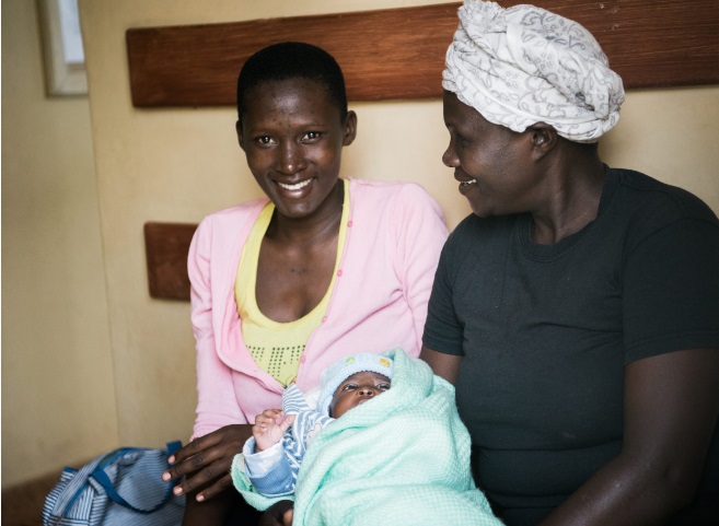 A birth companion carrying a newborn baby sits beside a new mother in a hospital