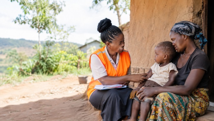 A community health worker in a bright orange vest visiting a rural household in East Africa, speaking to a mother with a young child. 