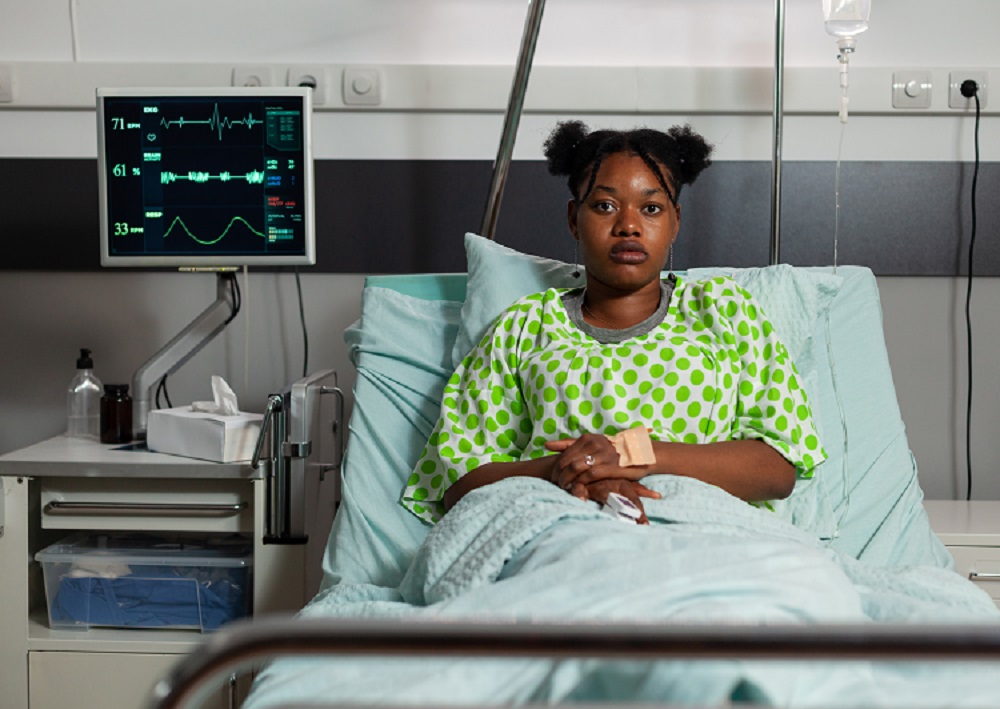 A young African woman in a green polka dot gown resting in a hospital bed and attached to a medical monitor and an IV drip. 