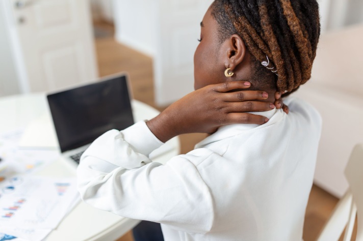 Back to side view of a young African lady sitting at a table and holding her neck in apparent pain while looking at a laptop screen.
