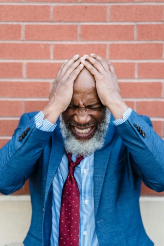 A distressed looking professioanl African male wearing a loose tie and blue jacket. He is holding his head presumably from eye strain induced headache.
