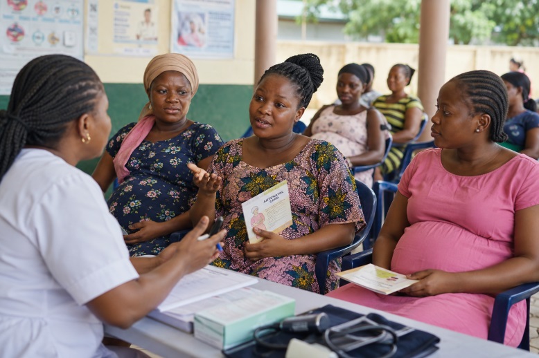 A pregnant African woman holding her antenatal card and sitting with others while speaking with a nurse during an antenatal visit.